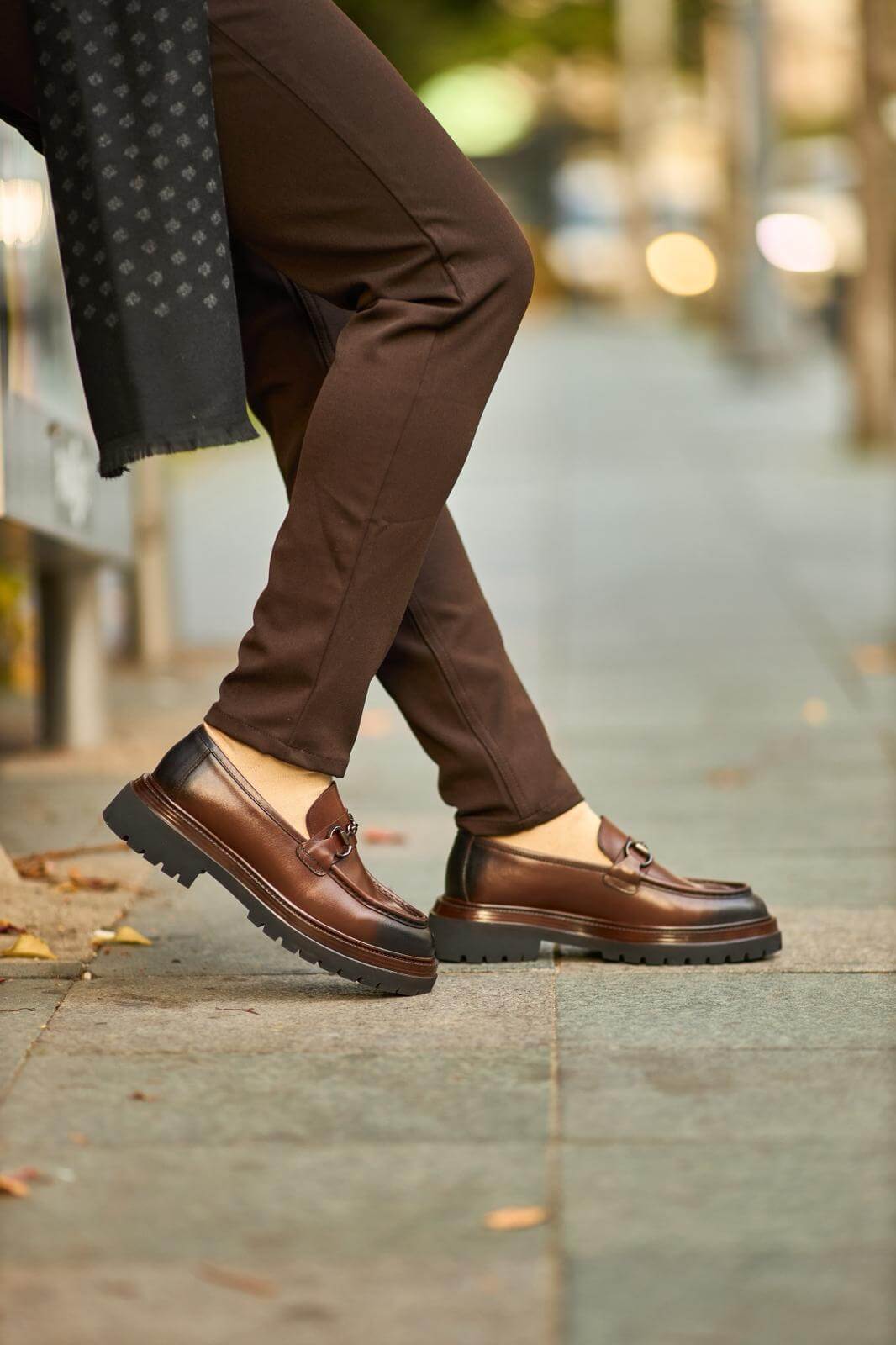 Brown leather bit loafers with chunky sole worn with brown trousers on outdoor pavement.