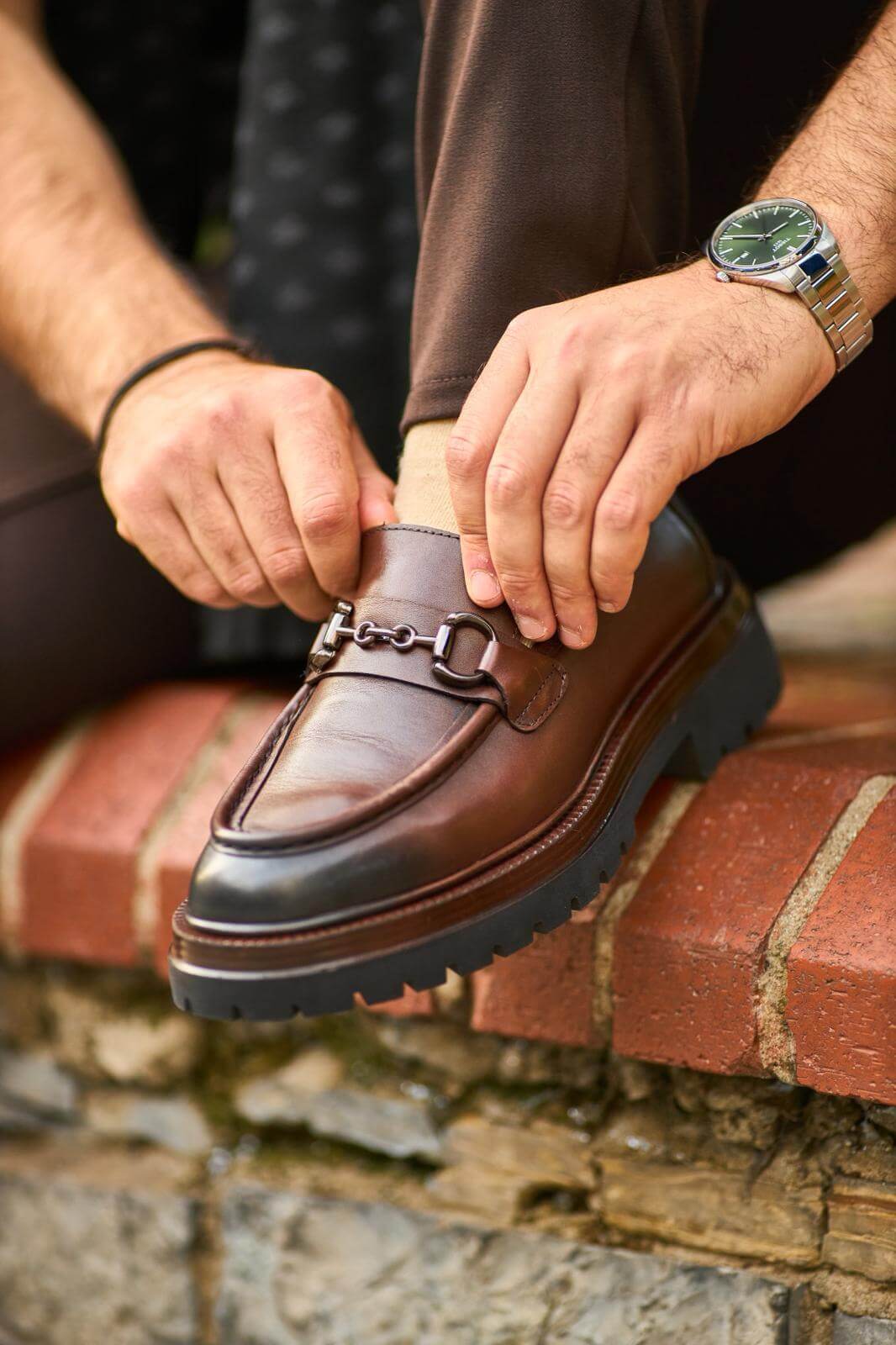 Brown leather bit loafers with chunky sole worn with brown trousers on outdoor pavement.