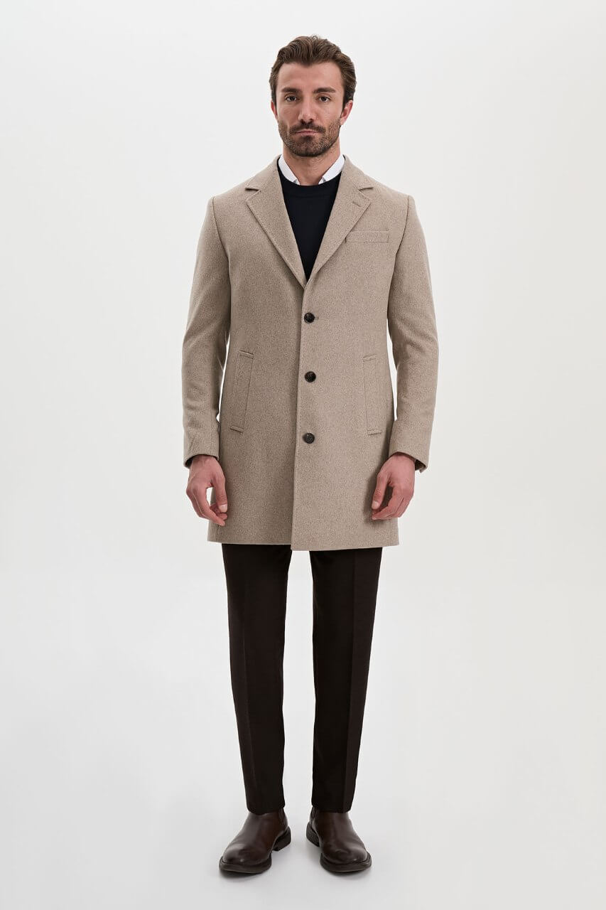 Men's beige single-breasted winter coat styled with a black sweater, dark trousers, and brown leather boots.