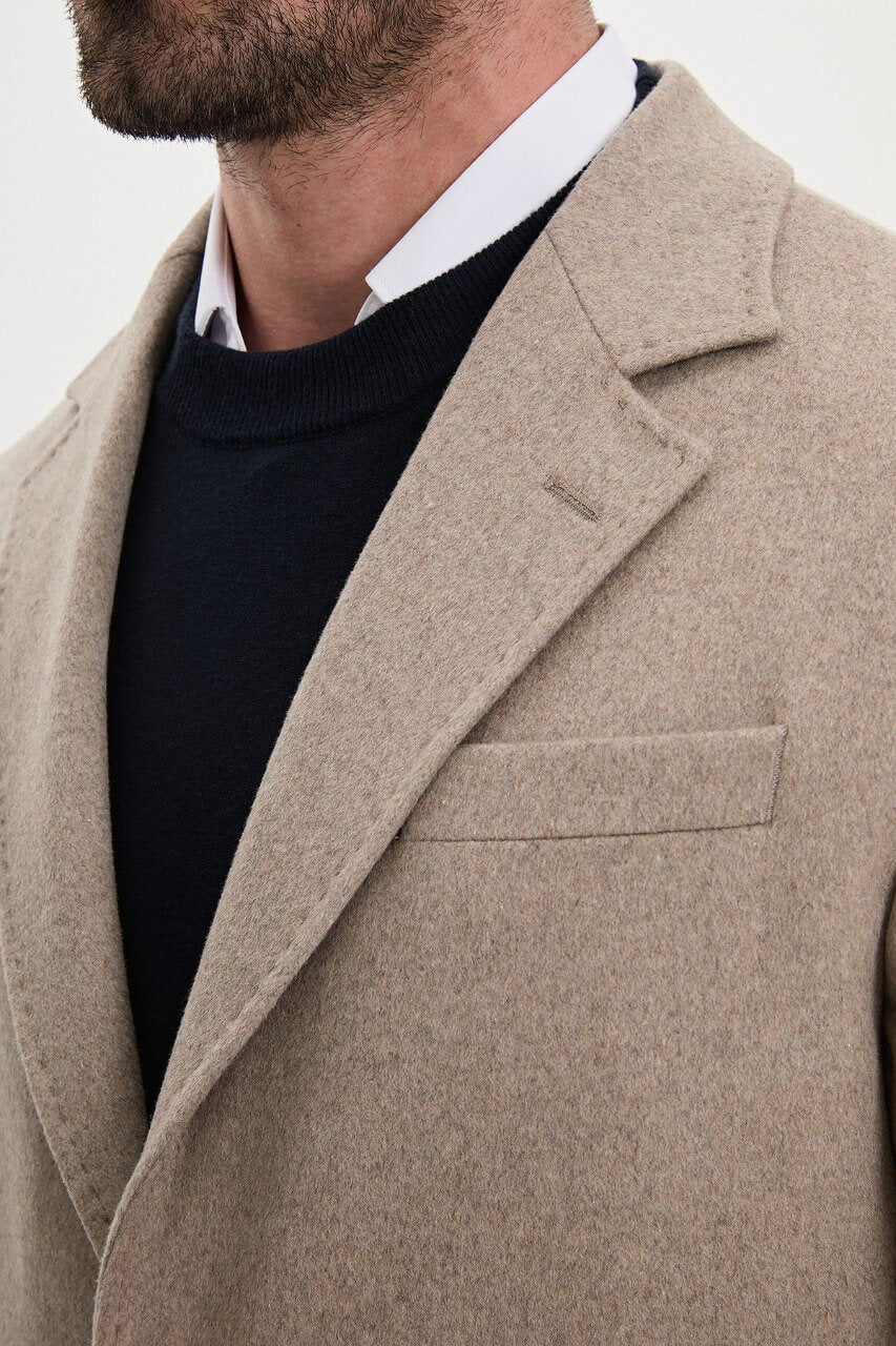 Men's beige single-breasted winter coat styled with a black sweater, dark trousers, and brown leather boots.