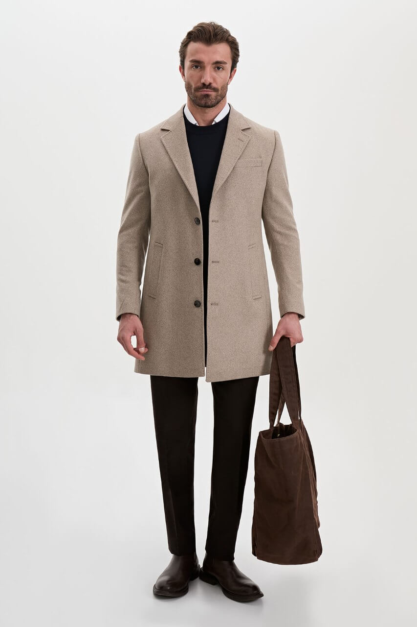 Men's beige single-breasted winter coat styled with a black sweater, dark trousers, and brown leather boots.