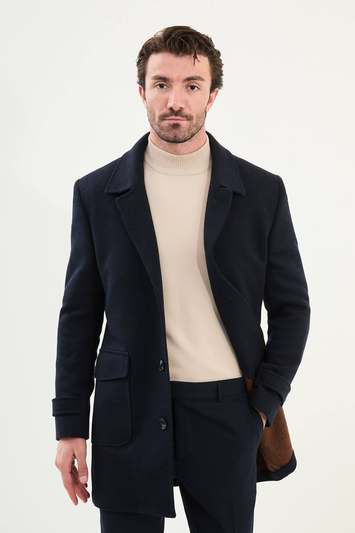 A man wearing a dark blue slim fit single-breasted coat over a beige turtleneck, styled with dark trousers and black shoes.