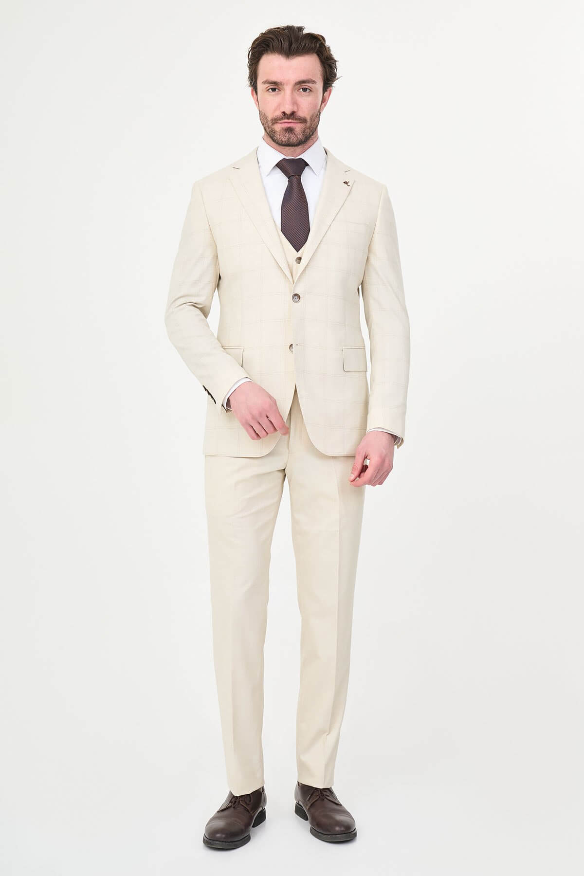 an ecru check slim-fit three-piece suit with matching waistcoat and trousers, white dress shirt, dark brown tie, and brown leather Derby shoes.