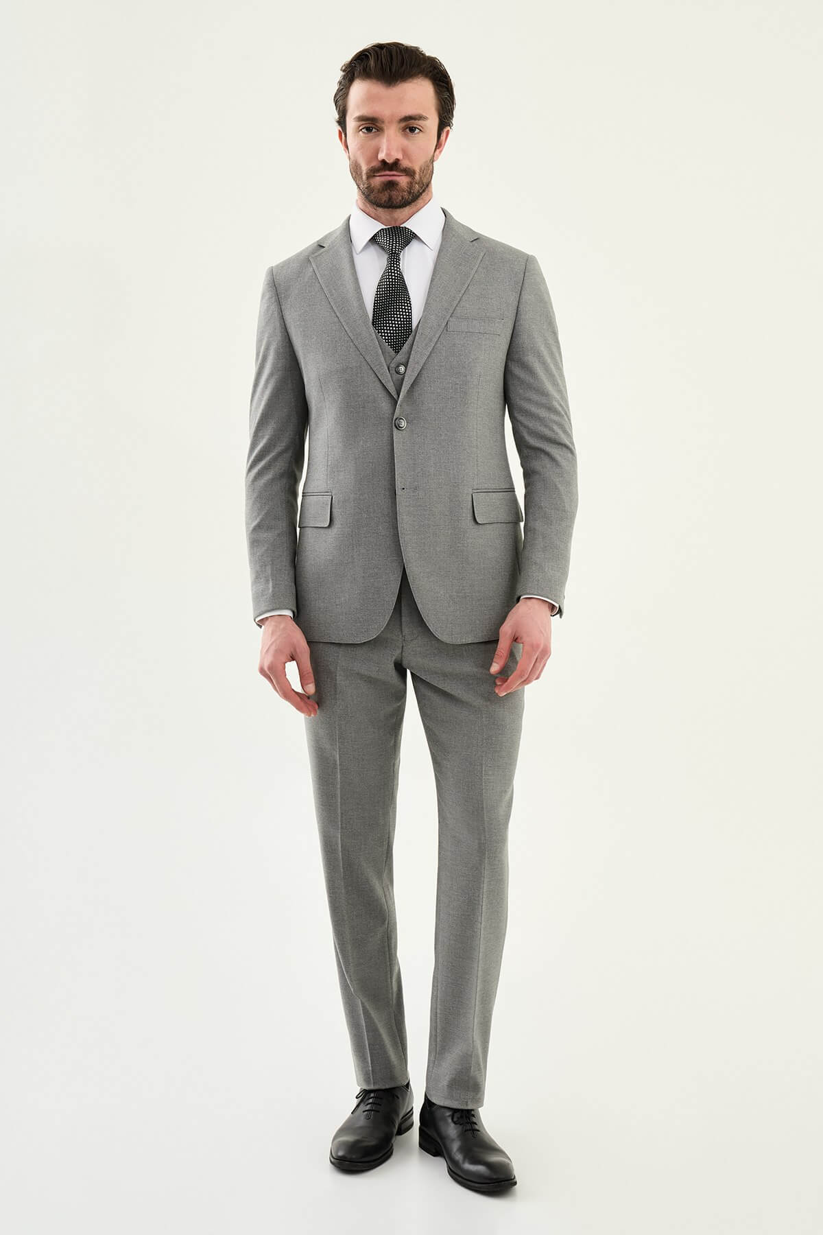 Man wearing a gray three-piece slim fit suit, including jacket, vest, and trousers, paired with a white dress shirt and black patterned tie