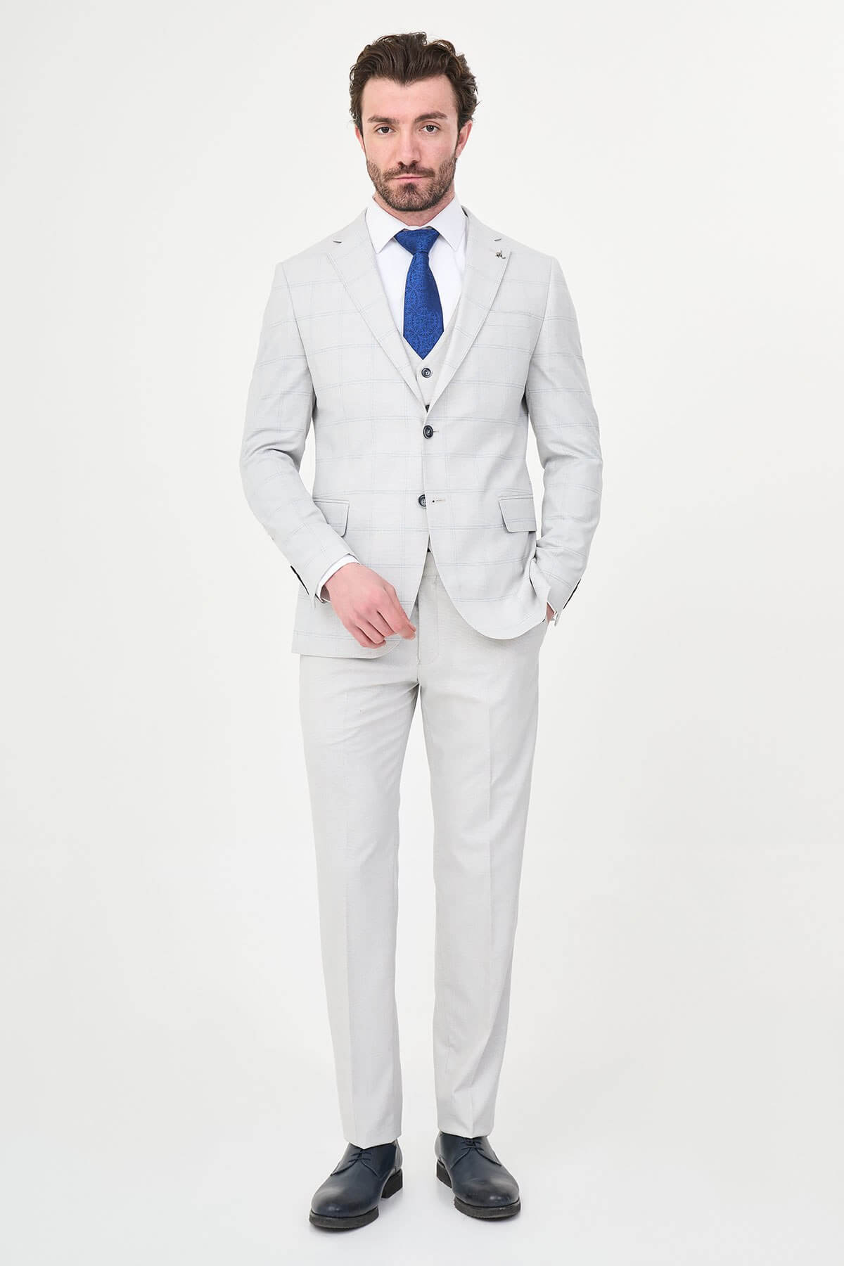 a gray check slim-fit three-piece suit with matching waistcoat and trousers, white dress shirt, royal-blue tie, and black leather Derby shoes.