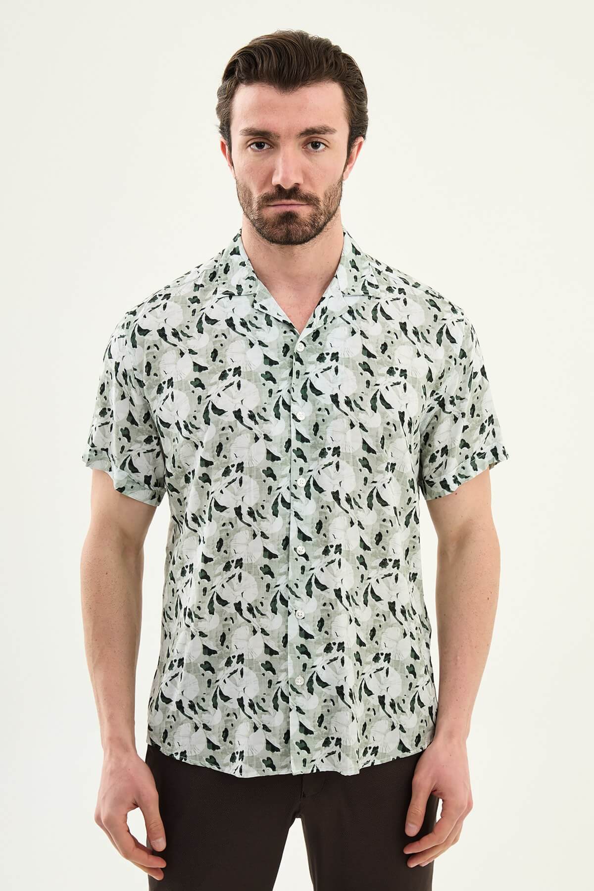Men wearing a green patterned short-sleeve summer shirt with tropical floral and leaf print, styled casually with light summer trousers.