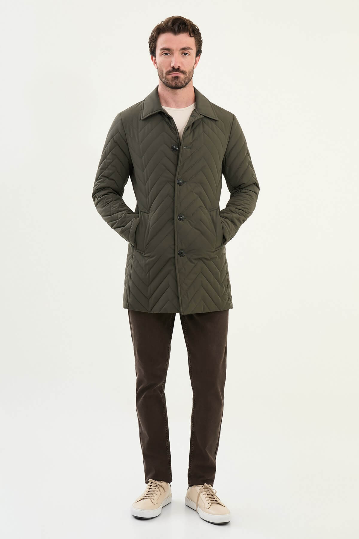 Man wearing a slim fit green quilted car coat with button closure, styled with brown trousers and beige sneakers.