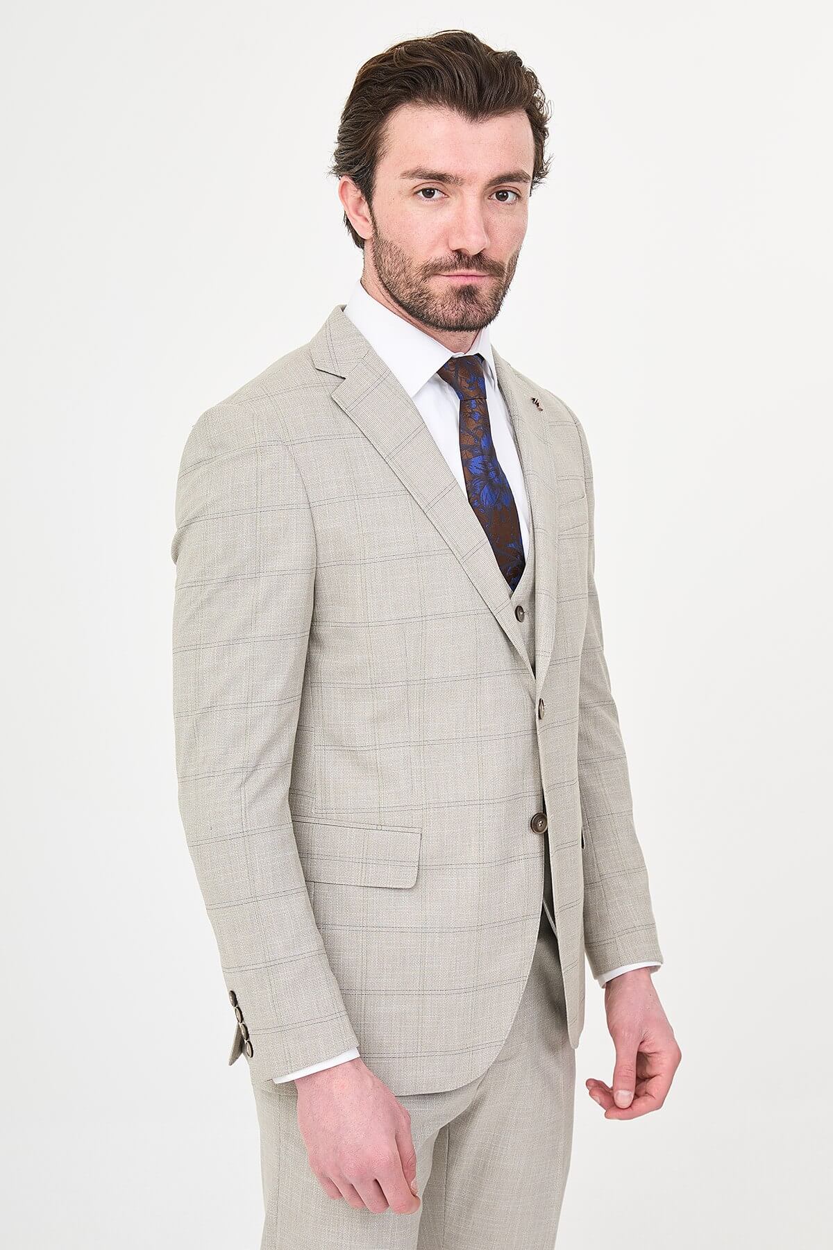 a mink slim-fit three-piece suit with a fine windowpane check, matching waistcoat and trousers, white dress shirt, patterned navy and brown tie, and dark brown leather shoes.