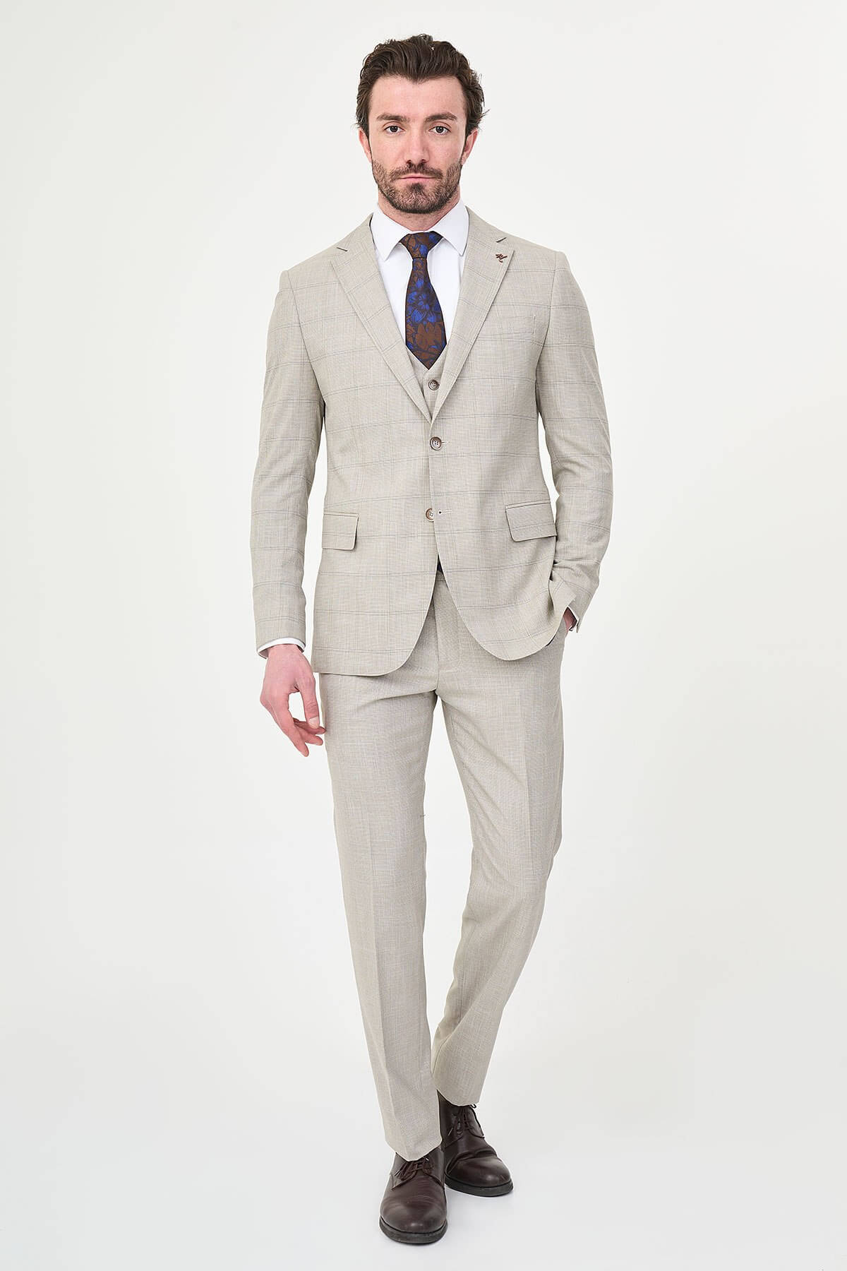 a mink slim-fit three-piece suit with a fine windowpane check, matching waistcoat and trousers, white dress shirt, patterned navy and brown tie, and dark brown leather shoes.