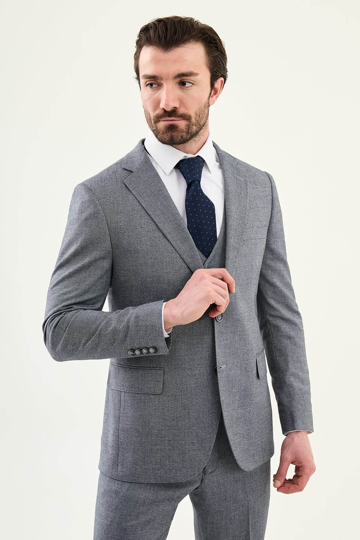 Model wearing a light grey slim-fit cotton-linen three-piece suit with a white dress shirt, navy dotted tie, and black Derby shoes.