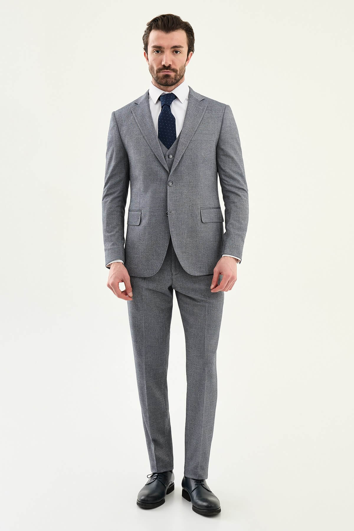 Model wearing a light grey slim-fit cotton-linen three-piece suit with a white dress shirt, navy dotted tie, and black Derby shoes.