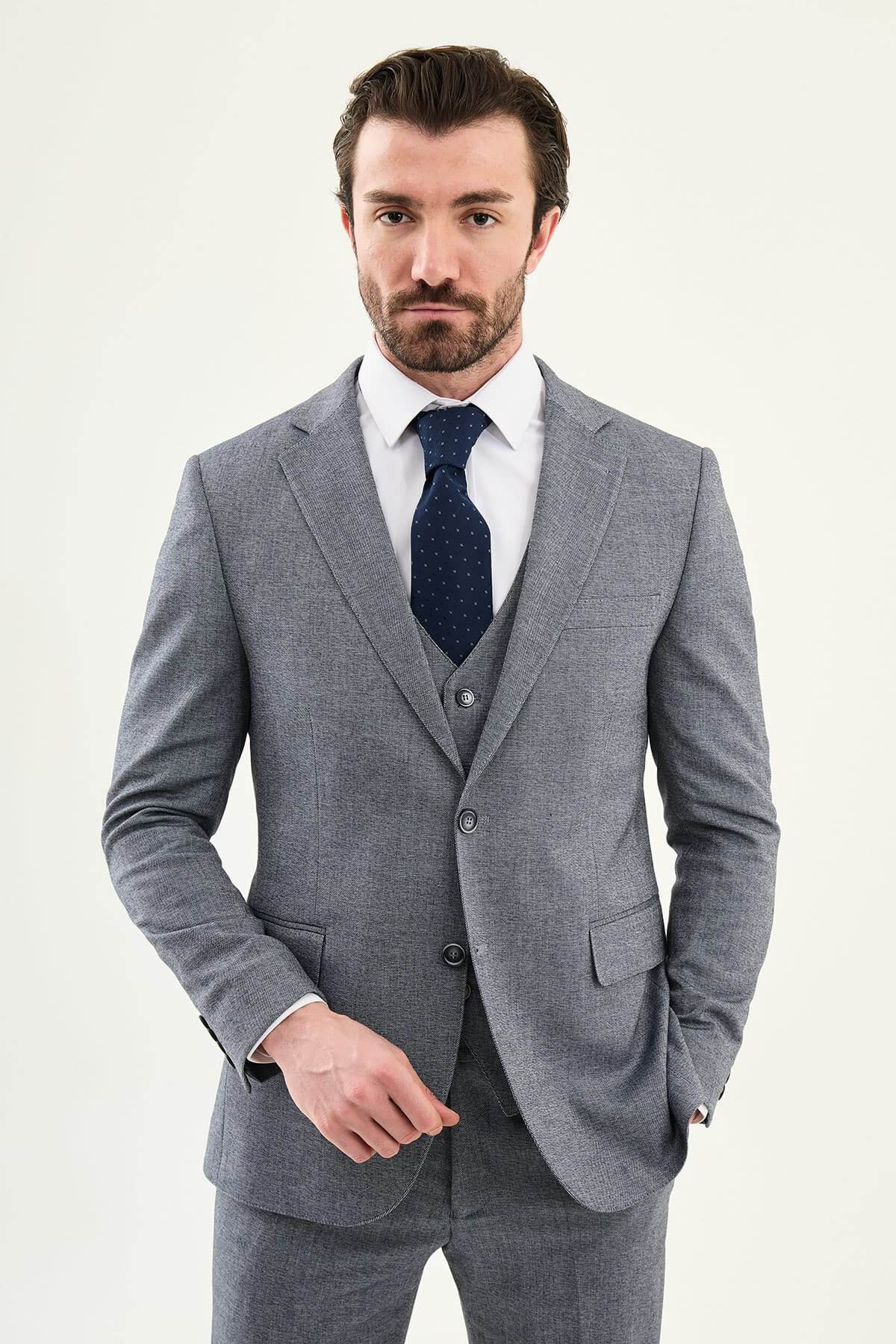 Model wearing a light grey slim-fit cotton-linen three-piece suit with a white dress shirt, navy dotted tie, and black Derby shoes.