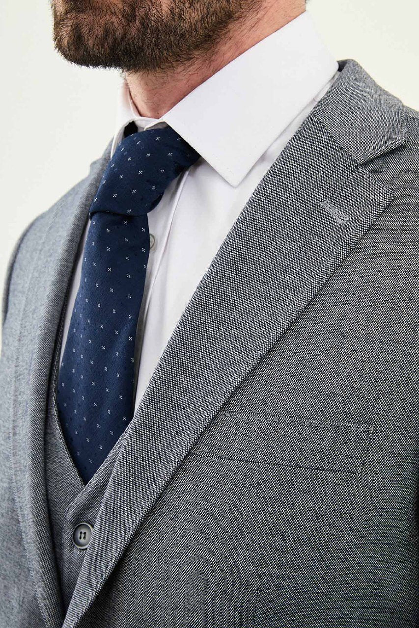Model wearing a light grey slim-fit cotton-linen three-piece suit with a white dress shirt, navy dotted tie, and black Derby shoes.