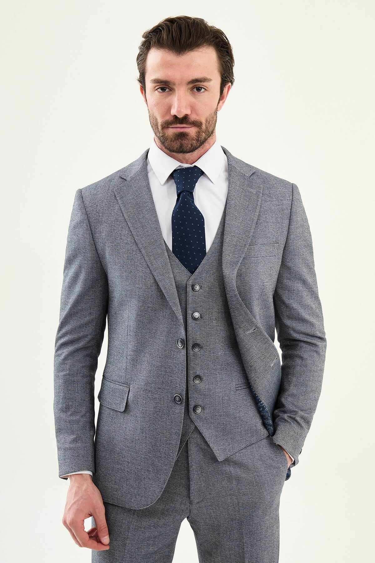 Model wearing a light grey slim-fit cotton-linen three-piece suit with a white dress shirt, navy dotted tie, and black Derby shoes.