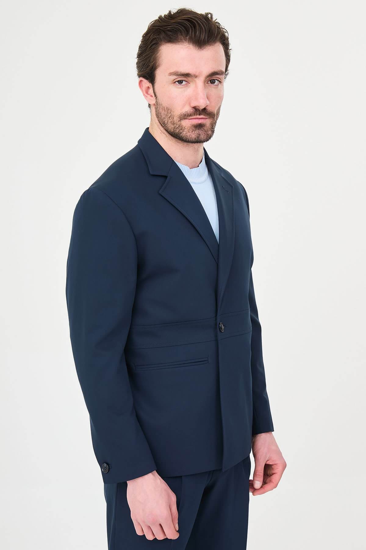 Man wearing a navy blue slim fit suit with black dress shoes, standing in a minimalist studio background, showcasing formal and elegant business attire.