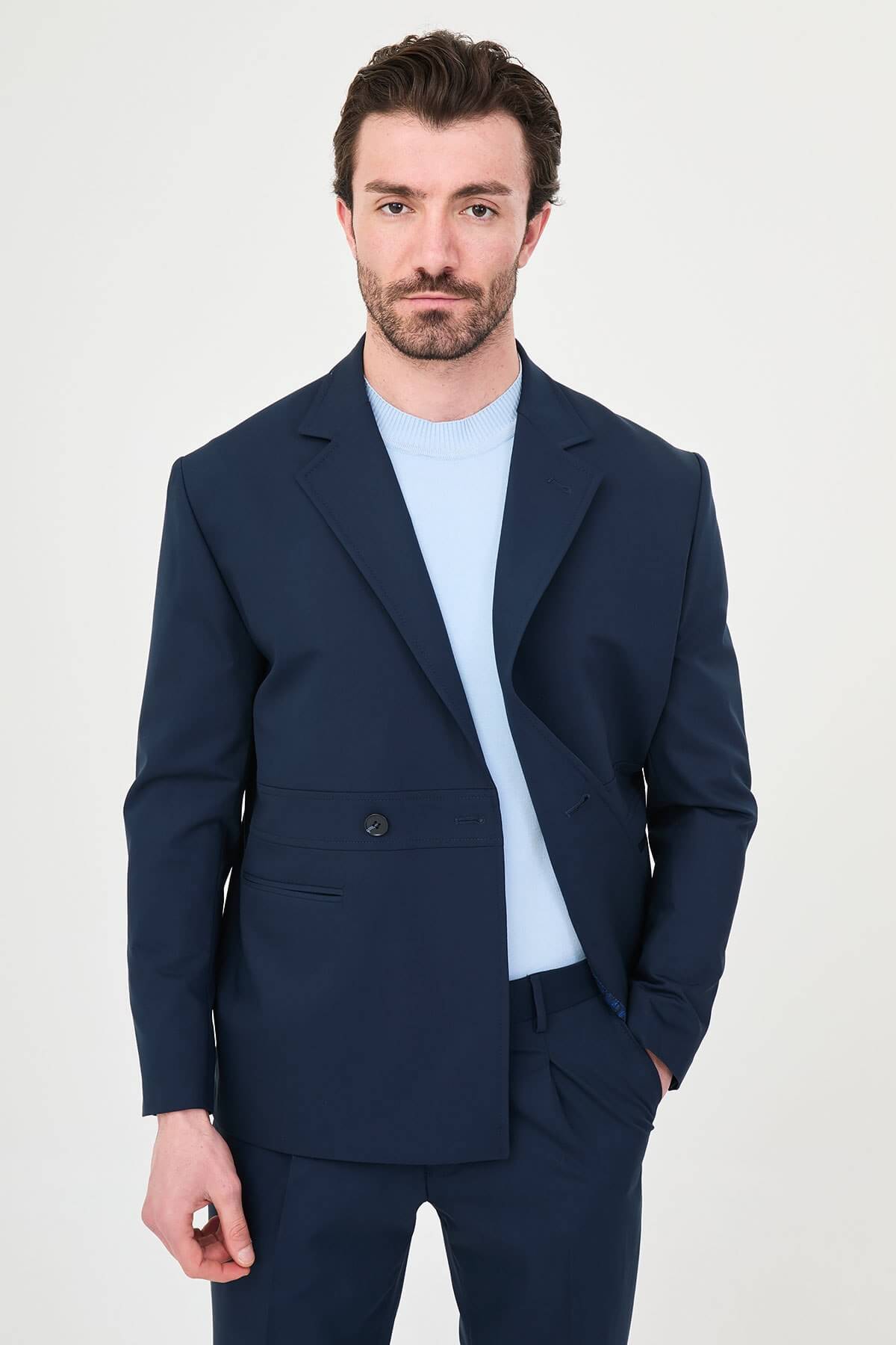 Man wearing a navy blue slim fit suit with black dress shoes, standing in a minimalist studio background, showcasing formal and elegant business attire.