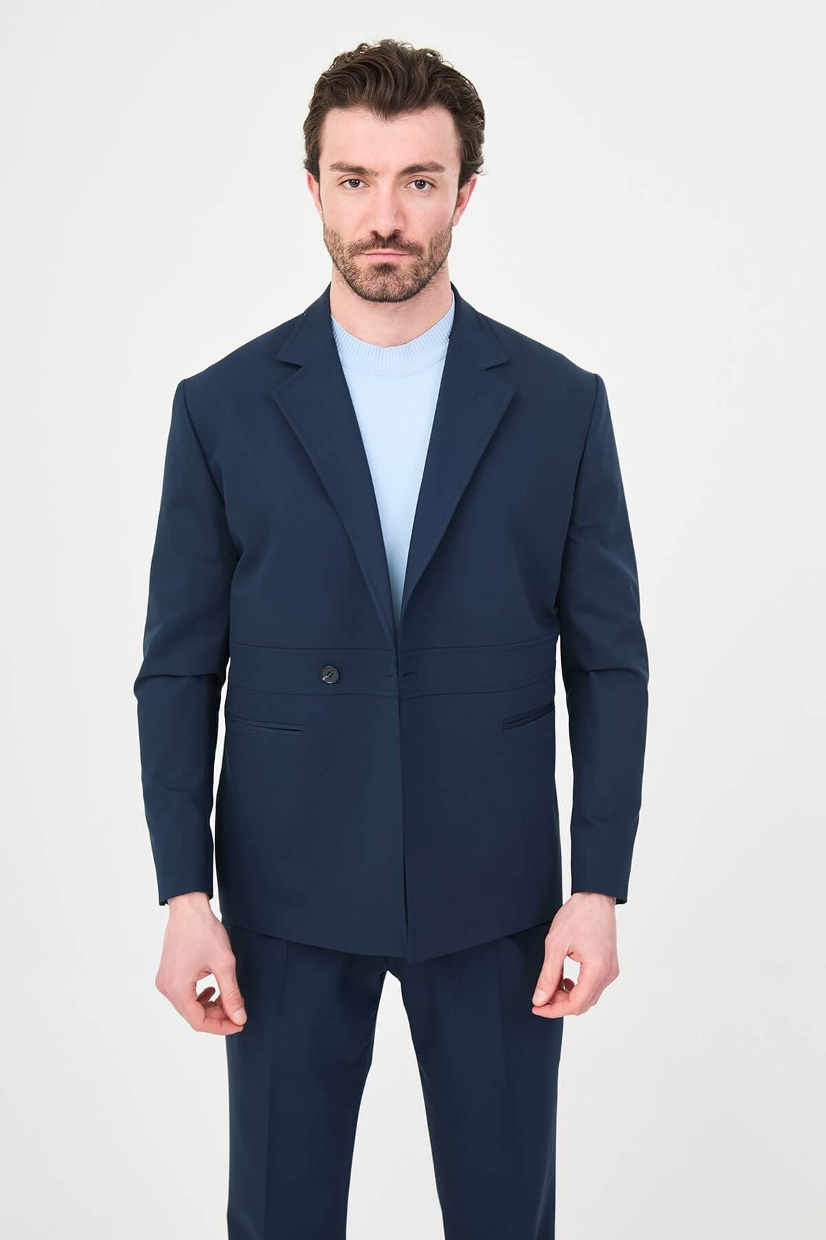 Man wearing a navy blue slim fit suit with black dress shoes, standing in a minimalist studio background, showcasing formal and elegant business attire.