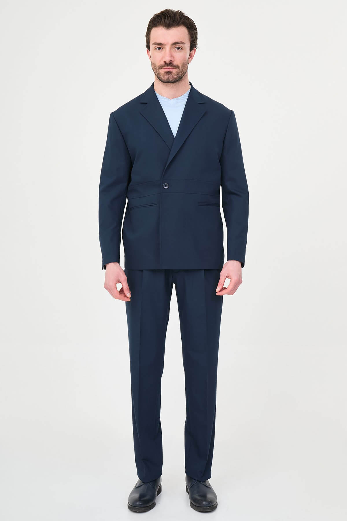 Man wearing a navy blue slim fit suit with black dress shoes, standing in a minimalist studio background, showcasing formal and elegant business attire.