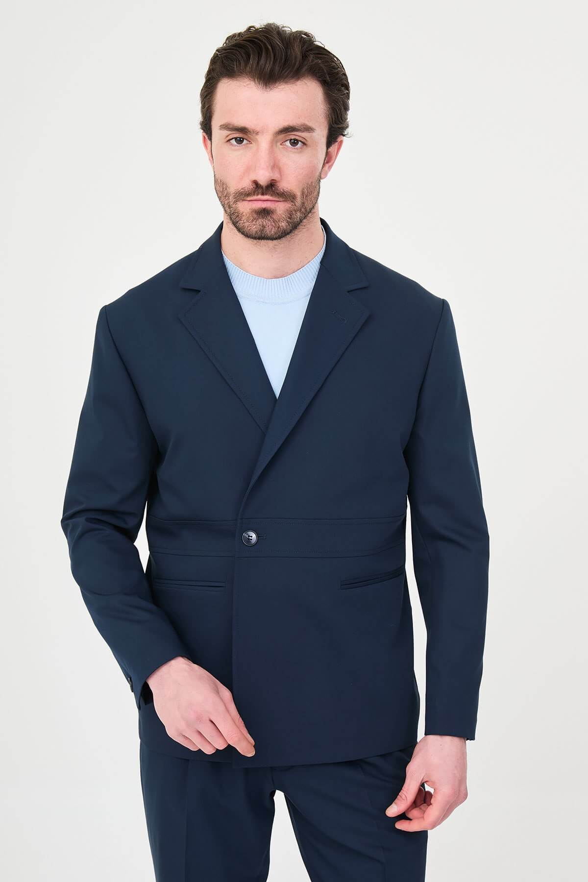 Man wearing a navy blue slim fit suit with black dress shoes, standing in a minimalist studio background, showcasing formal and elegant business attire.