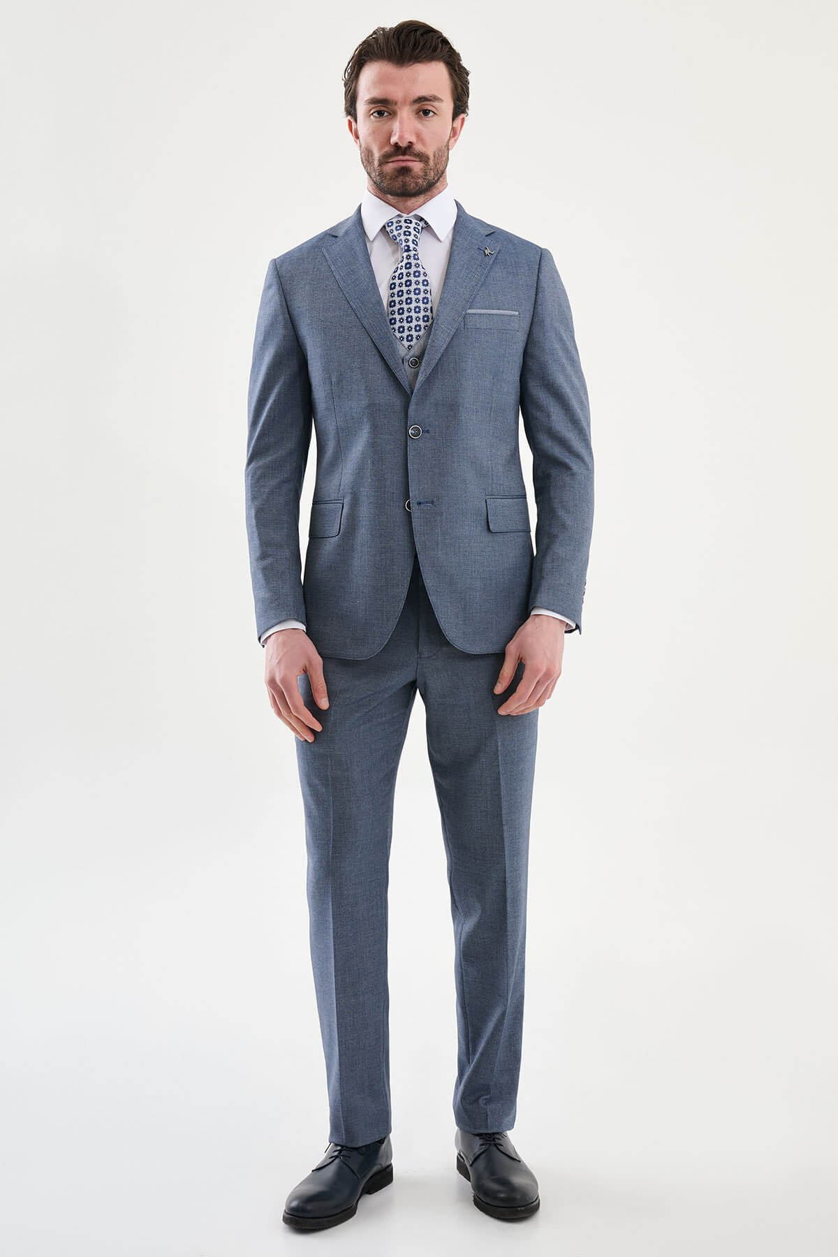 Model wearing a navy blue slim-fit three-piece suit with a white dress shirt, patterned blue tie, matching waistcoat, tailored trousers, and black leather dress shoes.