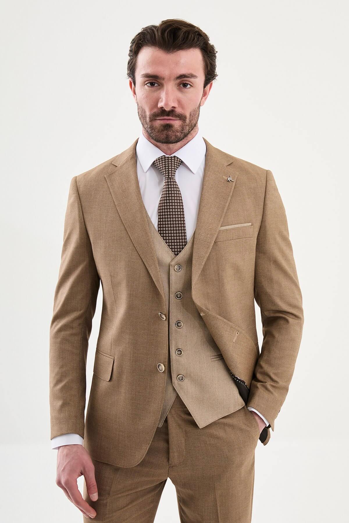 Model wearing a camel slim-fit three-piece suit with a white dress shirt, brown patterned tie, matching waistcoat, tailored trousers, and dark brown leather Derby shoes.