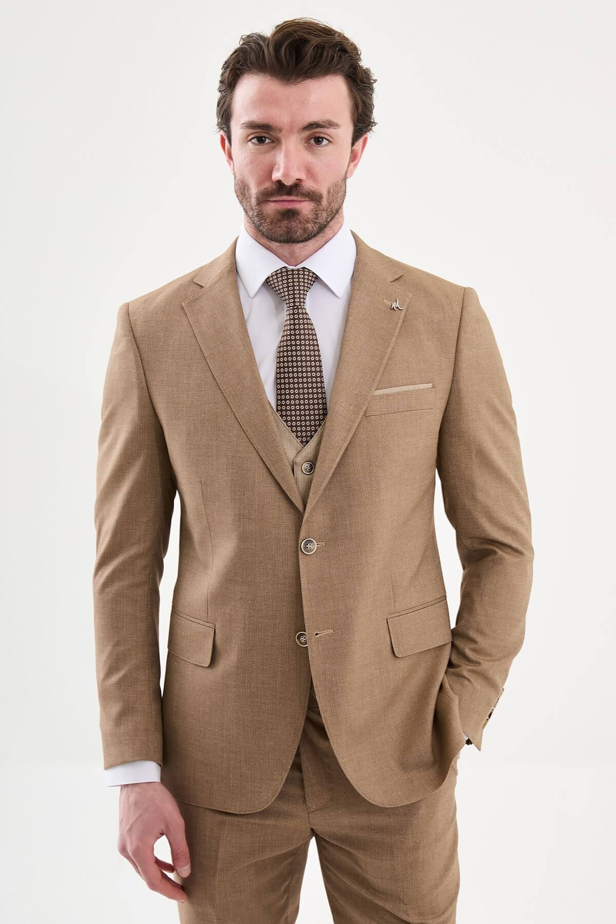 Model wearing a camel slim-fit three-piece suit with a white dress shirt, brown patterned tie, matching waistcoat, tailored trousers, and dark brown leather Derby shoes.