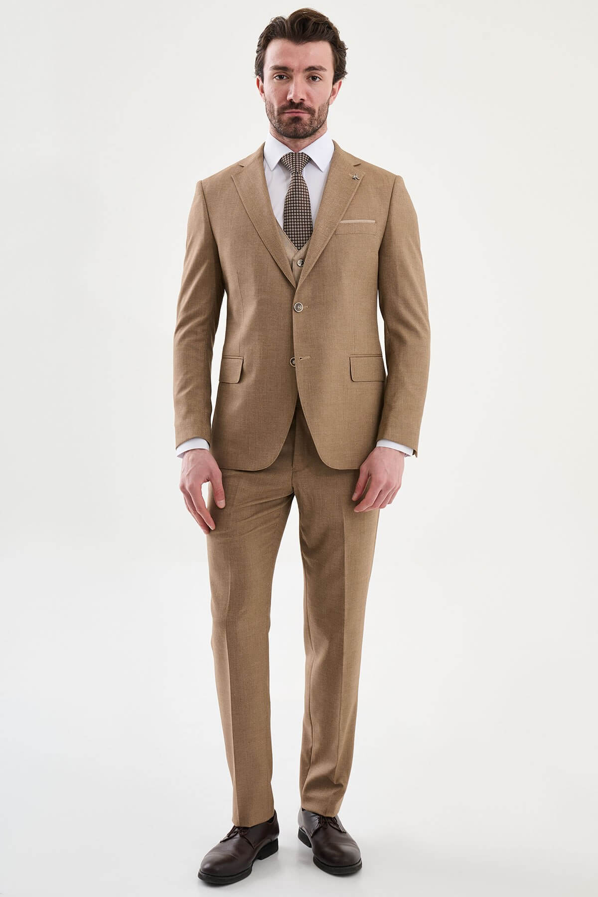 Model wearing a camel slim-fit three-piece suit with a white dress shirt, brown patterned tie, matching waistcoat, tailored trousers, and dark brown leather Derby shoes.