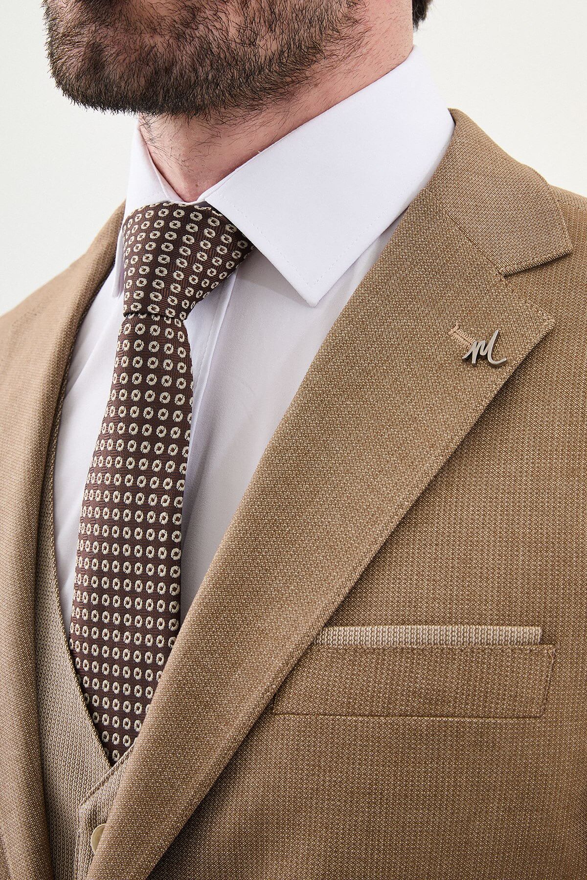 Model wearing a camel slim-fit three-piece suit with a white dress shirt, brown patterned tie, matching waistcoat, tailored trousers, and dark brown leather Derby shoes.