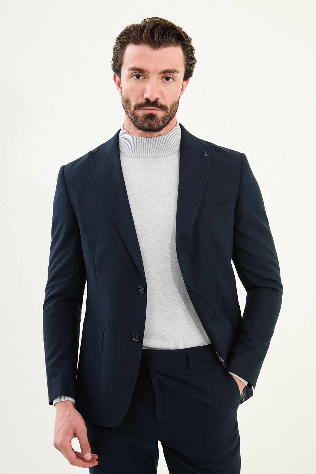 Men’s navy blue slim-fit suit with notch lapels and two-button jacket, matte micro-textured fabric, styled with a light gray mock-neck sweater for modern winter business wear.