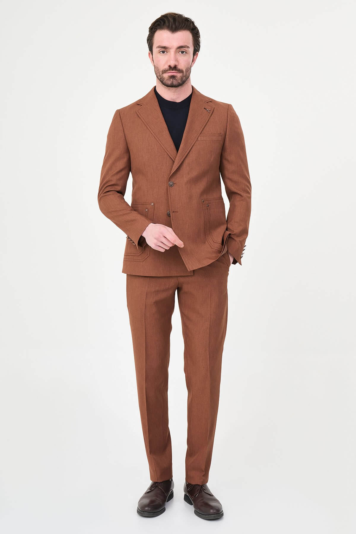 a tile slim-fit two-piece suit with notch lapels, utility-style stitched lower pockets, matching trousers, a black knit top, and brown leather shoes.