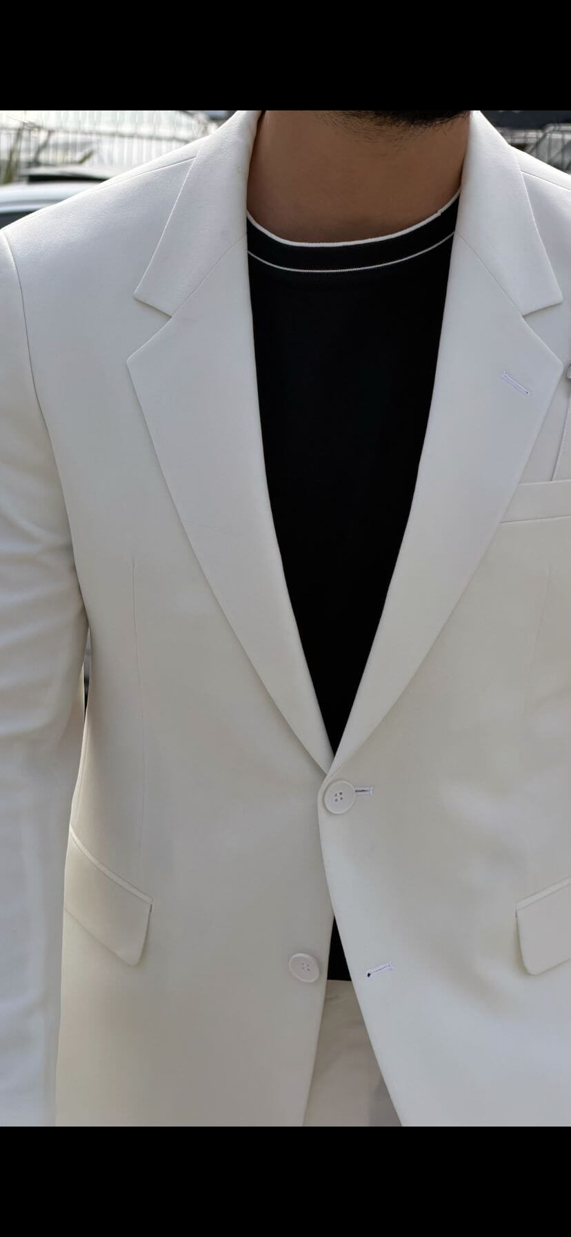 Man wearing a white shawl lapel slim-fit suit, styled with a black inner shirt, standing outdoors with yachts and a luxury car in the background.