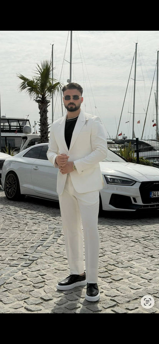 Man wearing a white shawl lapel slim-fit suit, styled with a black inner shirt, standing outdoors with yachts and a luxury car in the background.