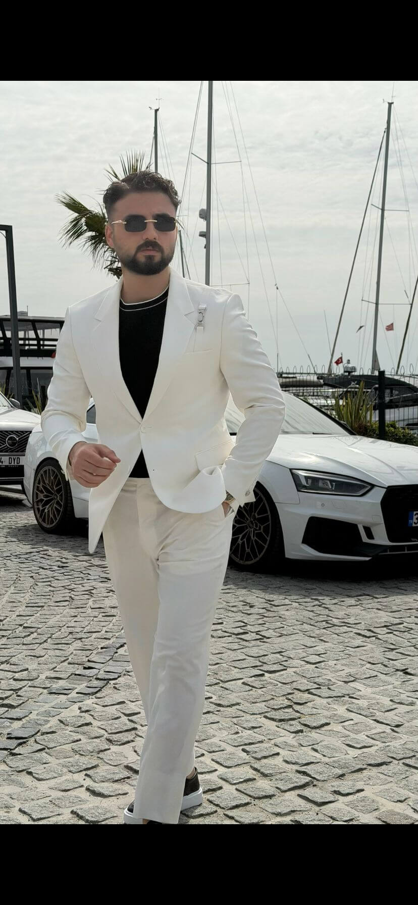 Man wearing a white shawl lapel slim-fit suit, styled with a black inner shirt, standing outdoors with yachts and a luxury car in the background.