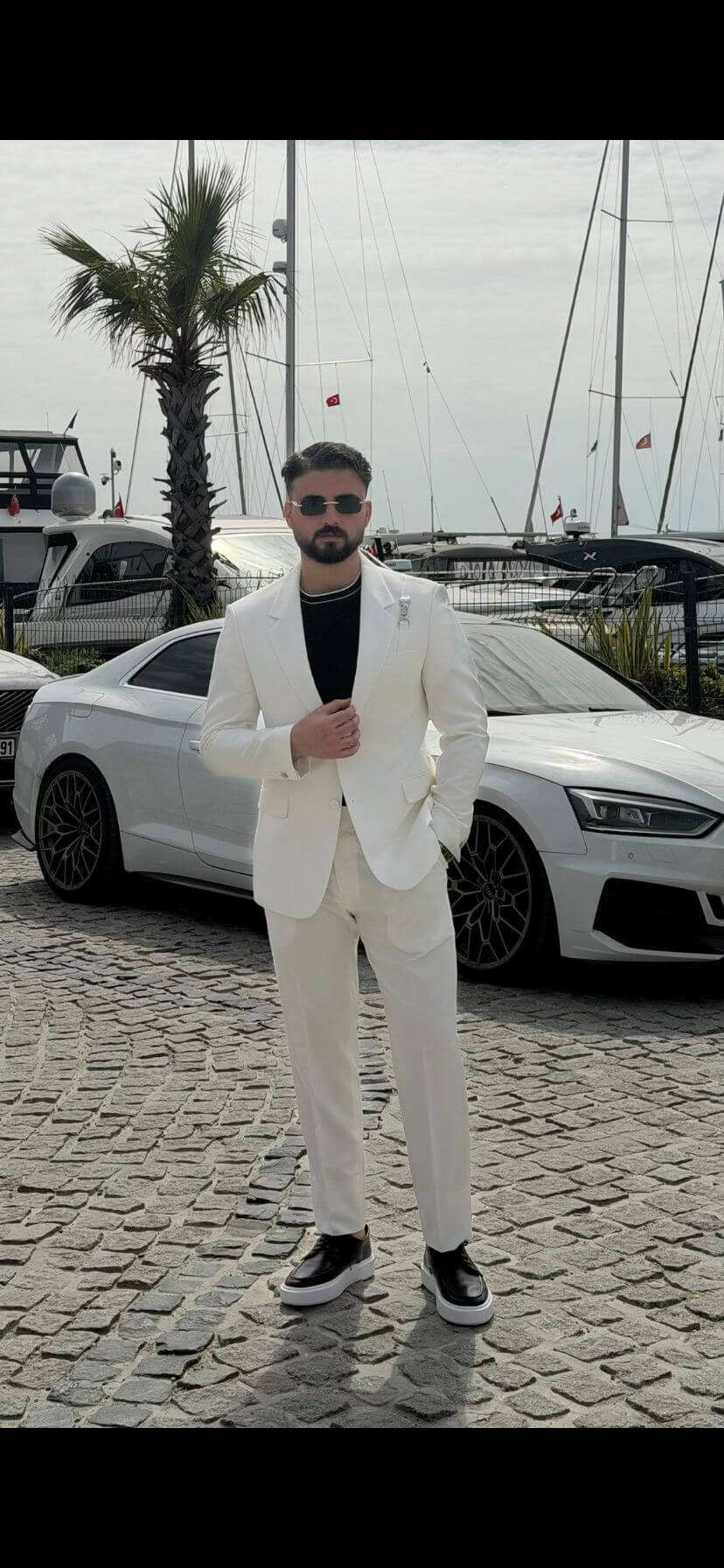 Man wearing a white shawl lapel slim-fit suit, styled with a black inner shirt, standing outdoors with yachts and a luxury car in the background.