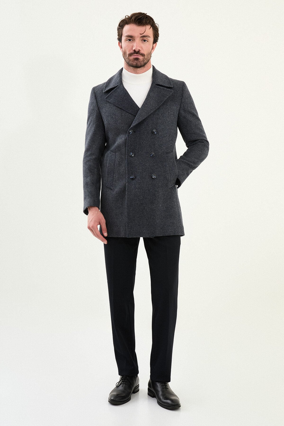Man wearing a dark gray slim-fit double-breasted peacoat over a white turtleneck with black trousers and black shoes.