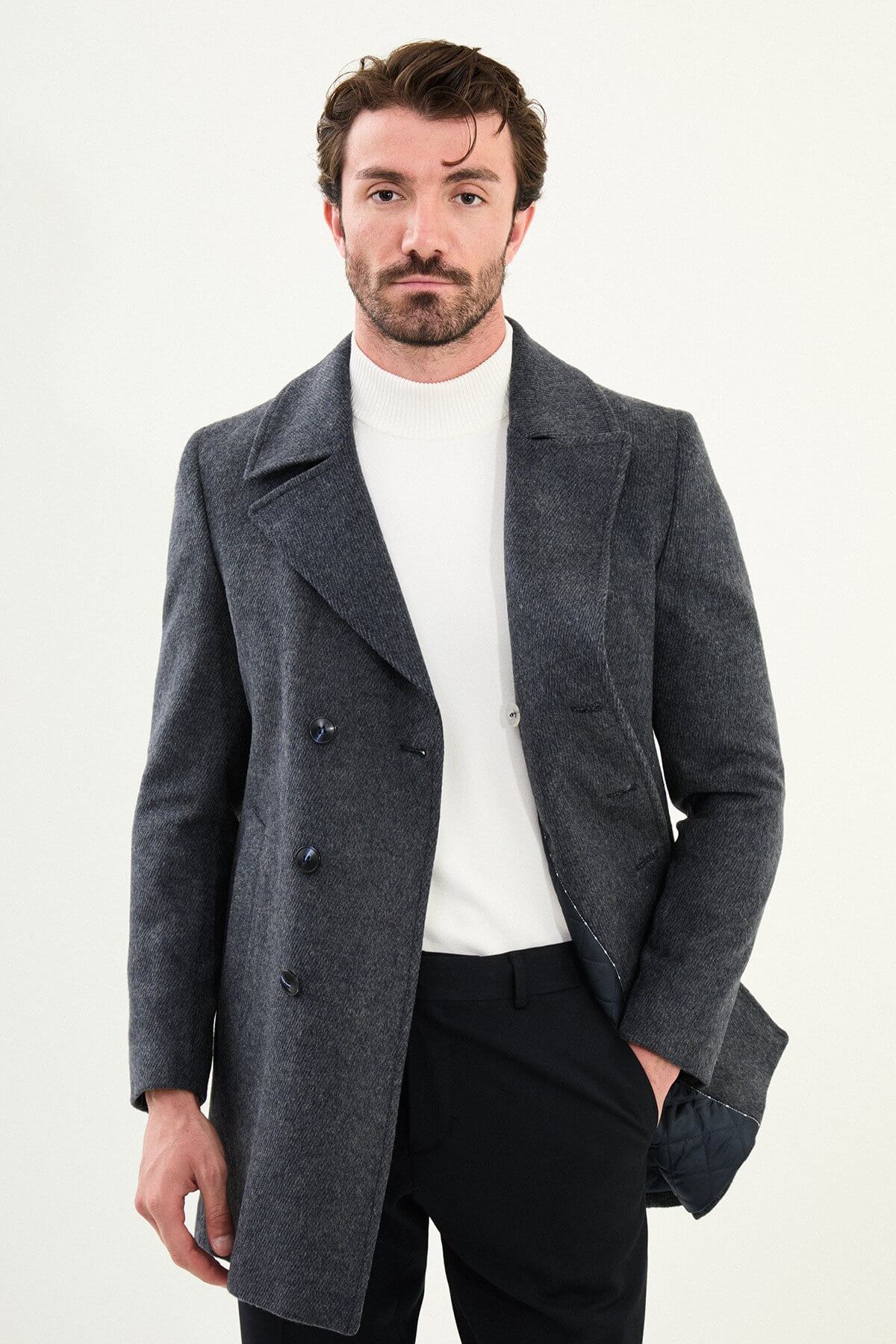Man wearing a dark gray slim-fit double-breasted peacoat over a white turtleneck with black trousers and black shoes.