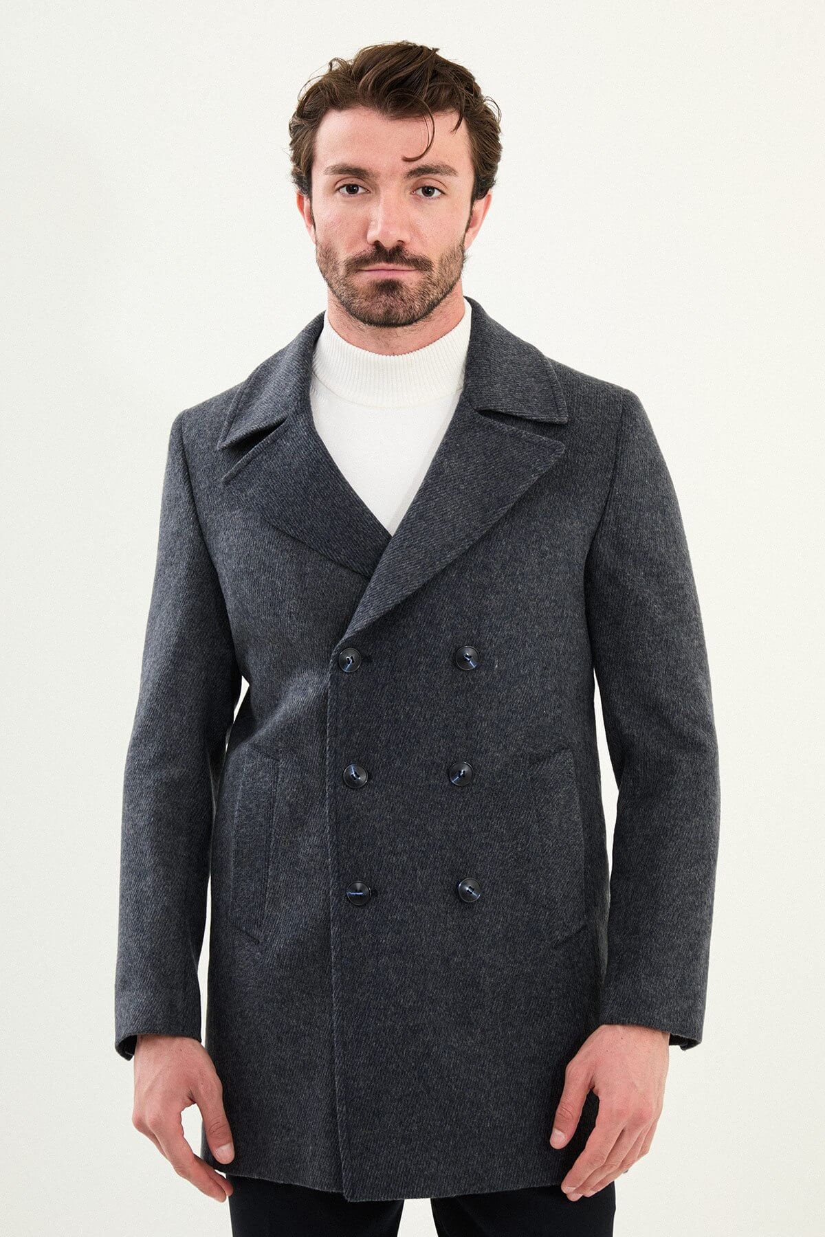 Man wearing a dark gray slim-fit double-breasted peacoat over a white turtleneck with black trousers and black shoes.