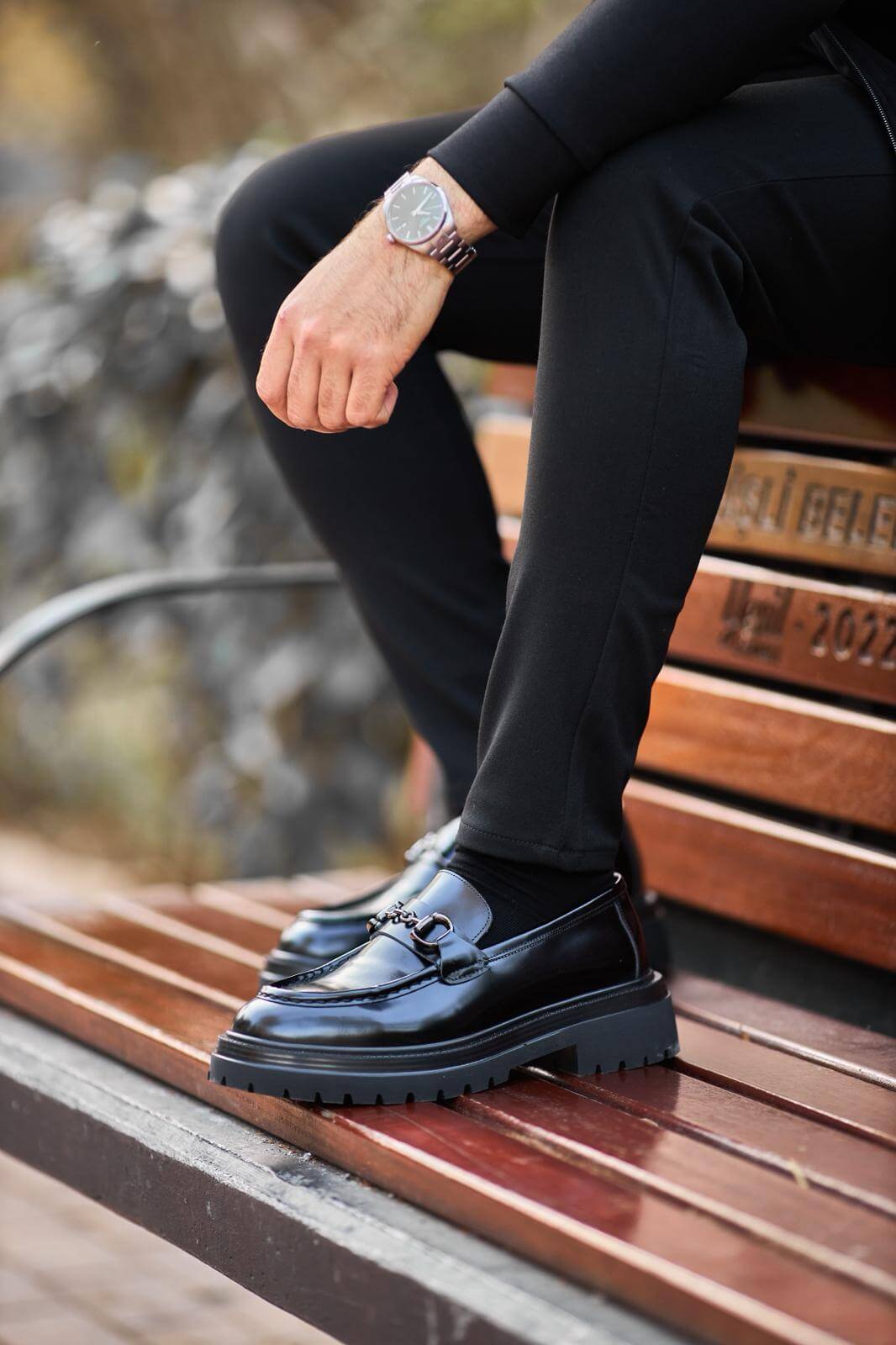 Man wearing glossy black leather horsebit loafers with chunky lug soles while sitting on a wooden bench.