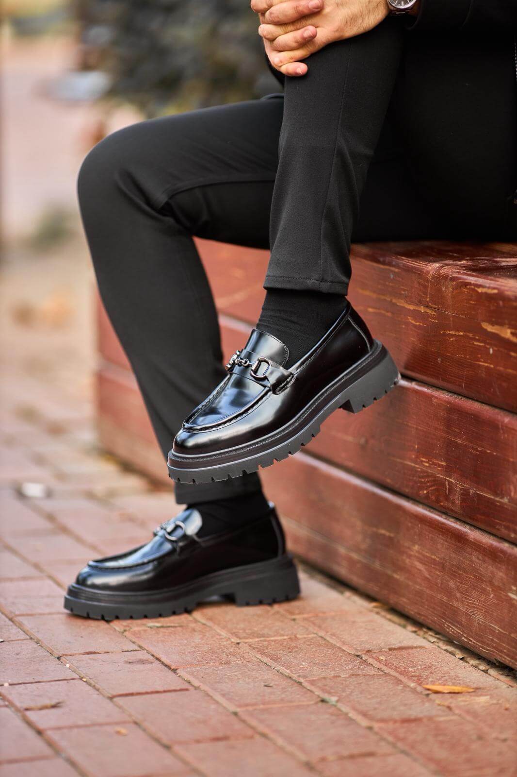 Man wearing glossy black leather horsebit loafers with chunky lug soles while sitting on a wooden bench.
