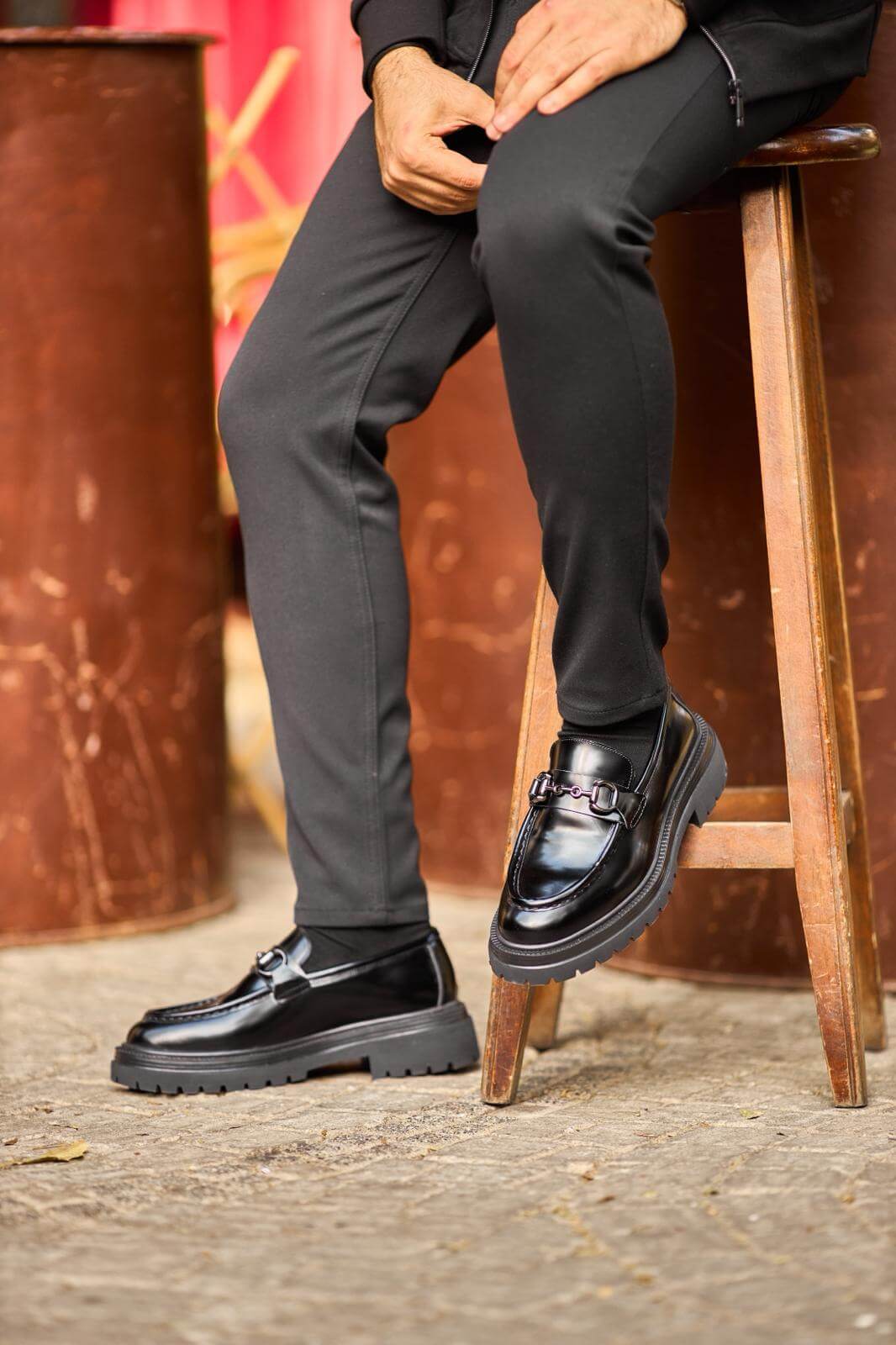 Man wearing glossy black leather horsebit loafers with chunky lug soles while sitting on a wooden bench.