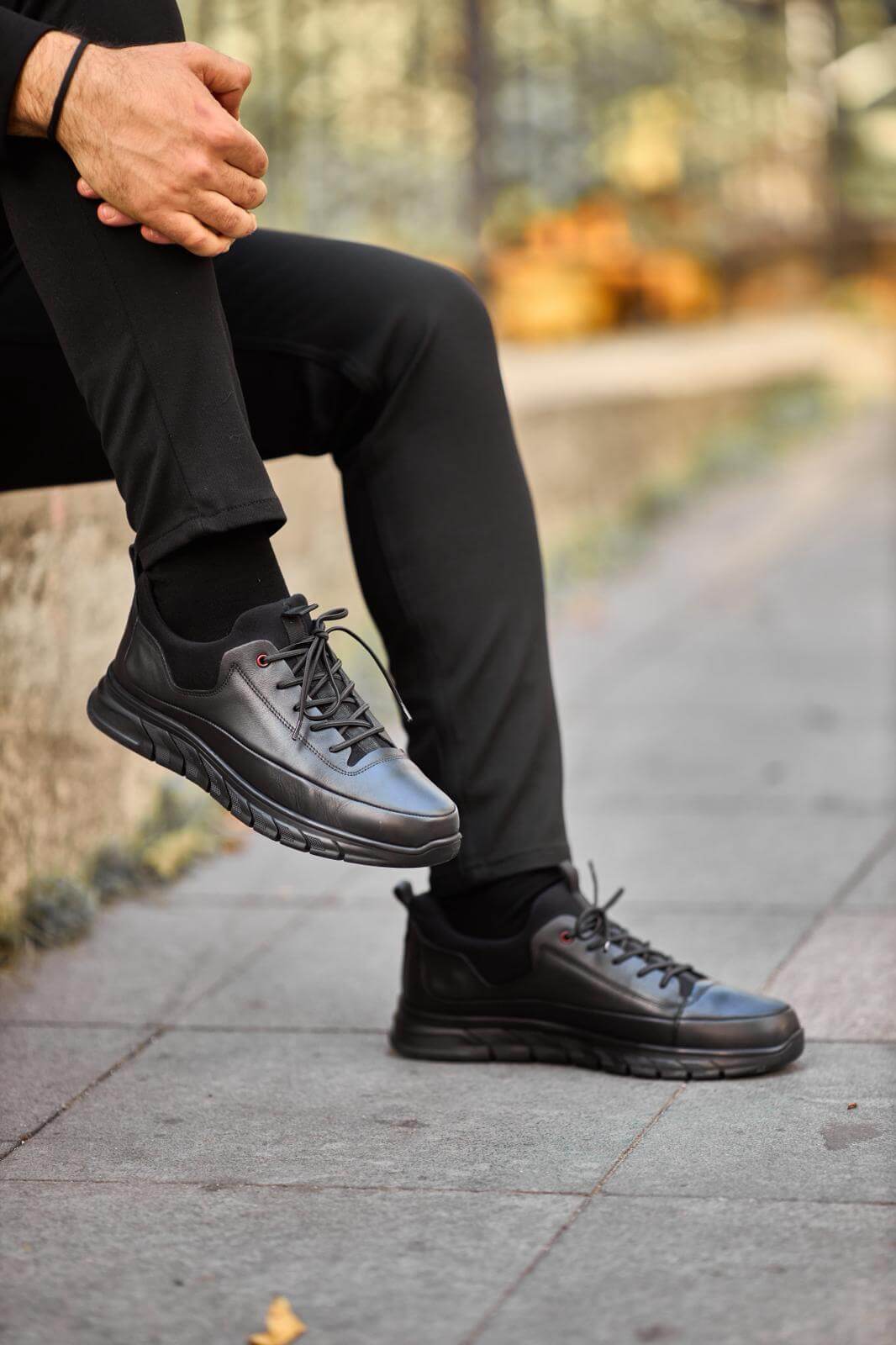 Black full-leather chunky-sole sneaker worn with black pants, photographed while the model sits on a concrete ledge outdoors.