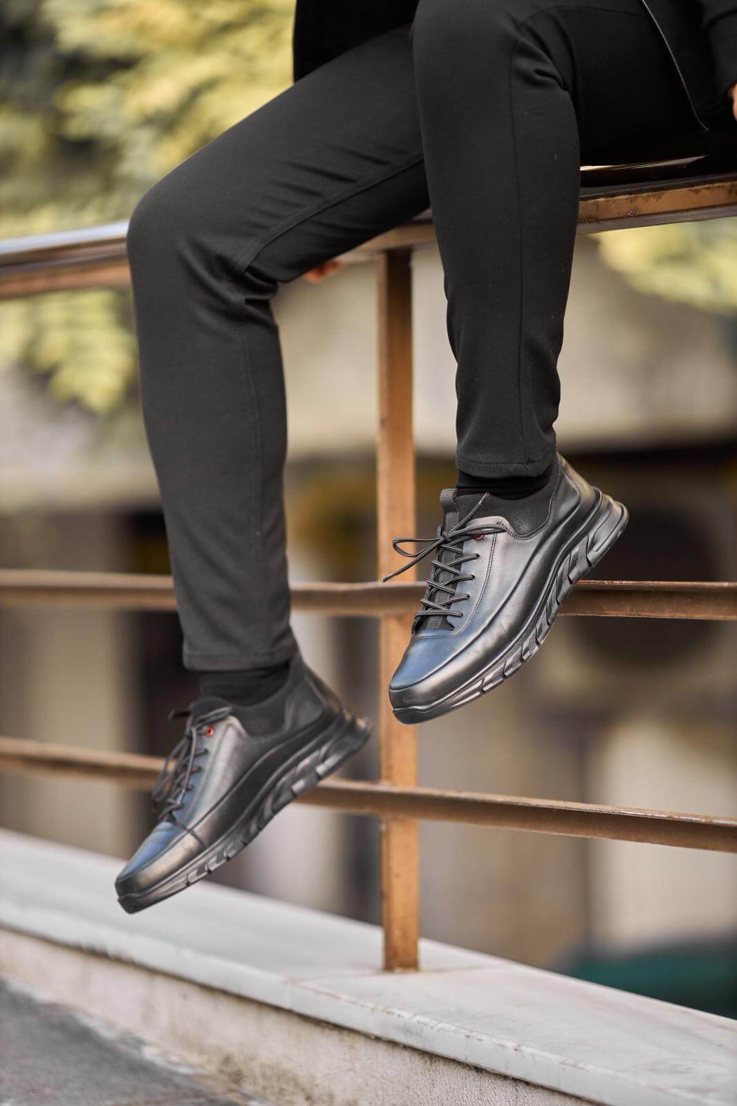 Black full-leather chunky-sole sneaker worn with black pants, photographed while the model sits on a concrete ledge outdoors.