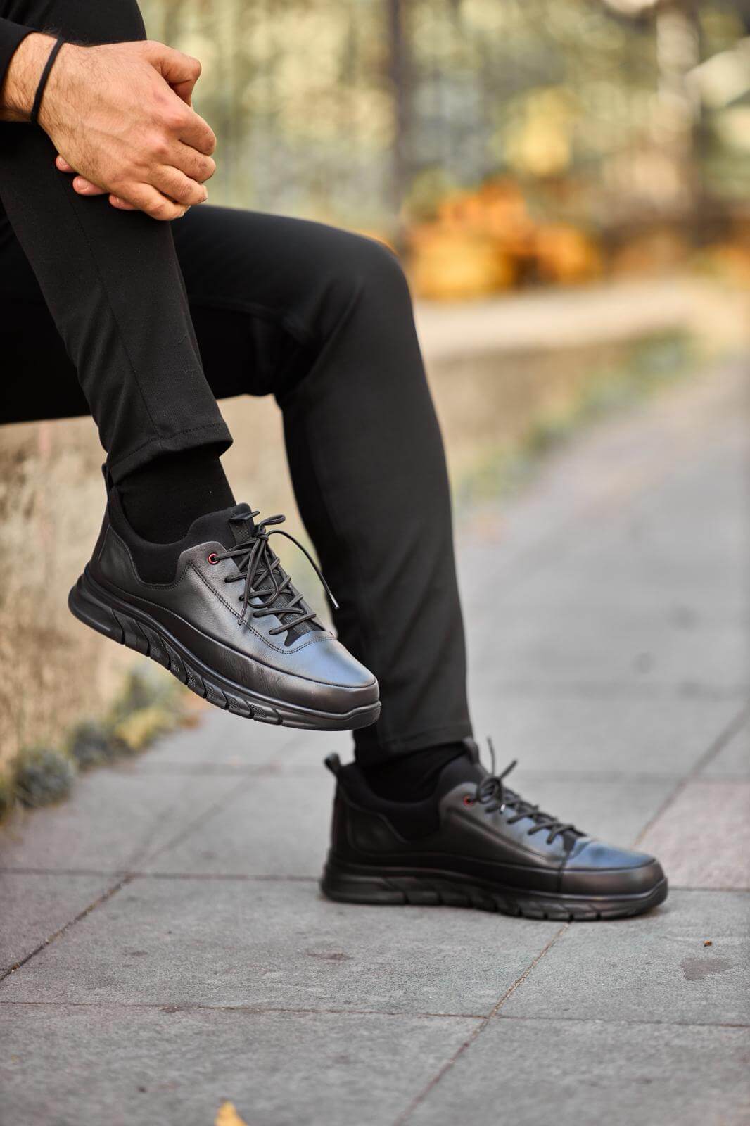 Black full-leather chunky-sole sneaker worn with black pants, photographed while the model sits on a concrete ledge outdoors.