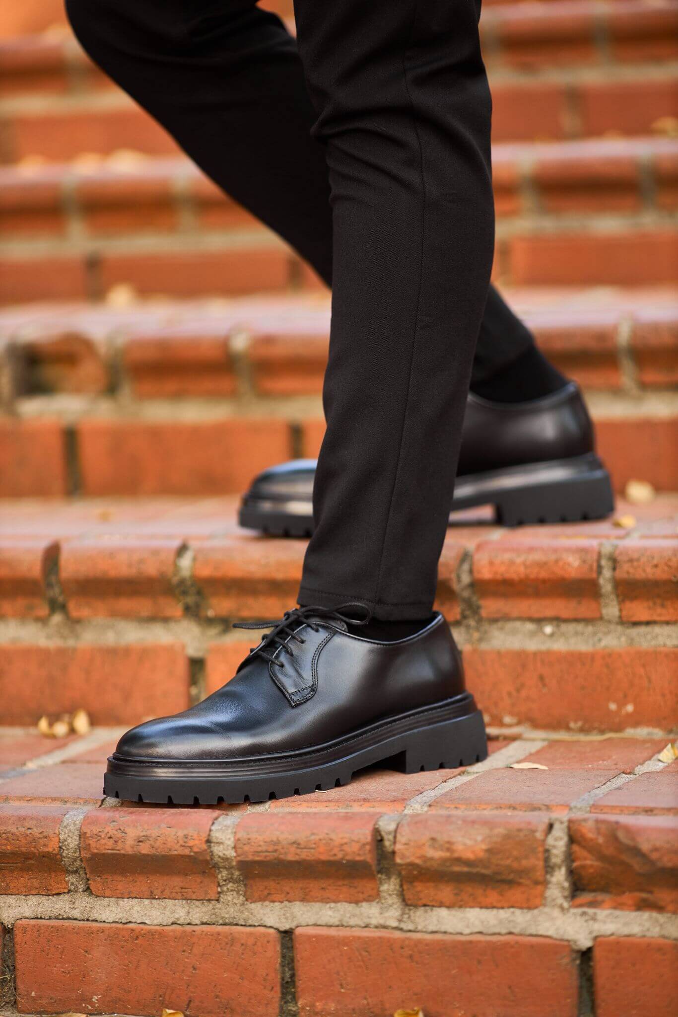  A man wearing black chunky-sole leather Derby shoes while walking down outdoor brick steps. 