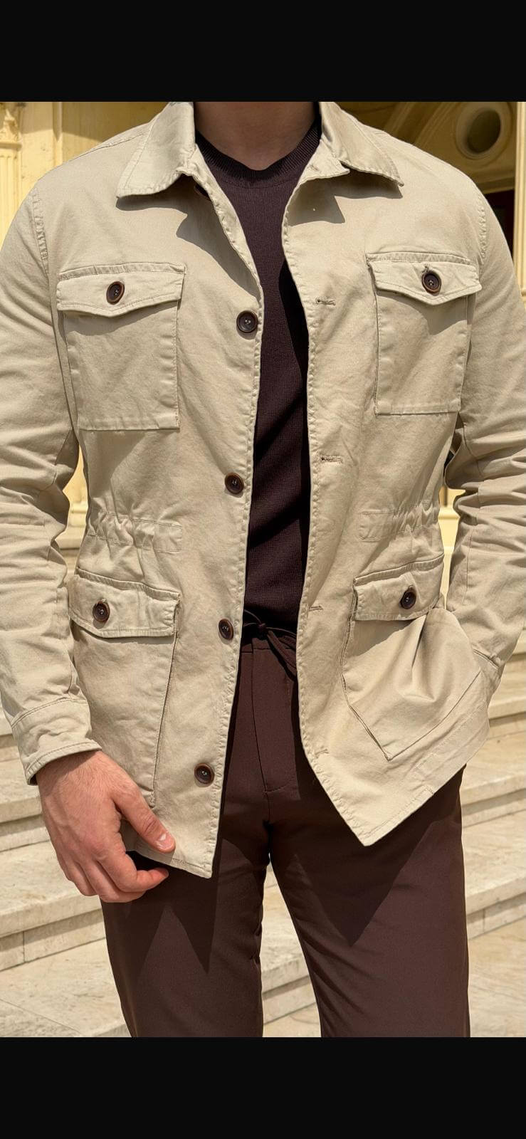 Men’s beige field jacket with multiple buttoned pockets, worn with brown trousers and a dark top, showcasing a casual outdoor look.