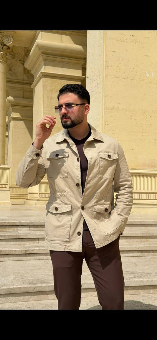 Men’s beige field jacket with multiple buttoned pockets, worn with brown trousers and a dark top, showcasing a casual outdoor look.