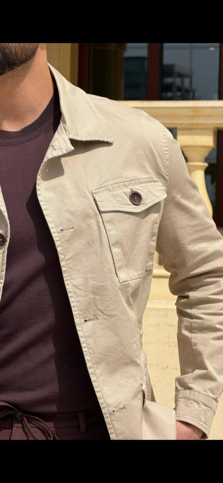Men’s beige field jacket with multiple buttoned pockets, worn with brown trousers and a dark top, showcasing a casual outdoor look.