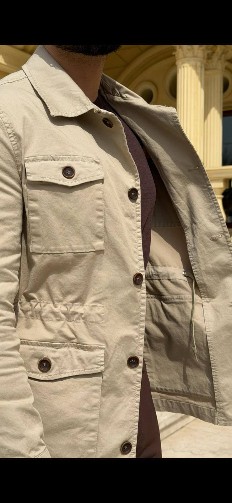 Men’s beige field jacket with multiple buttoned pockets, worn with brown trousers and a dark top, showcasing a casual outdoor look.