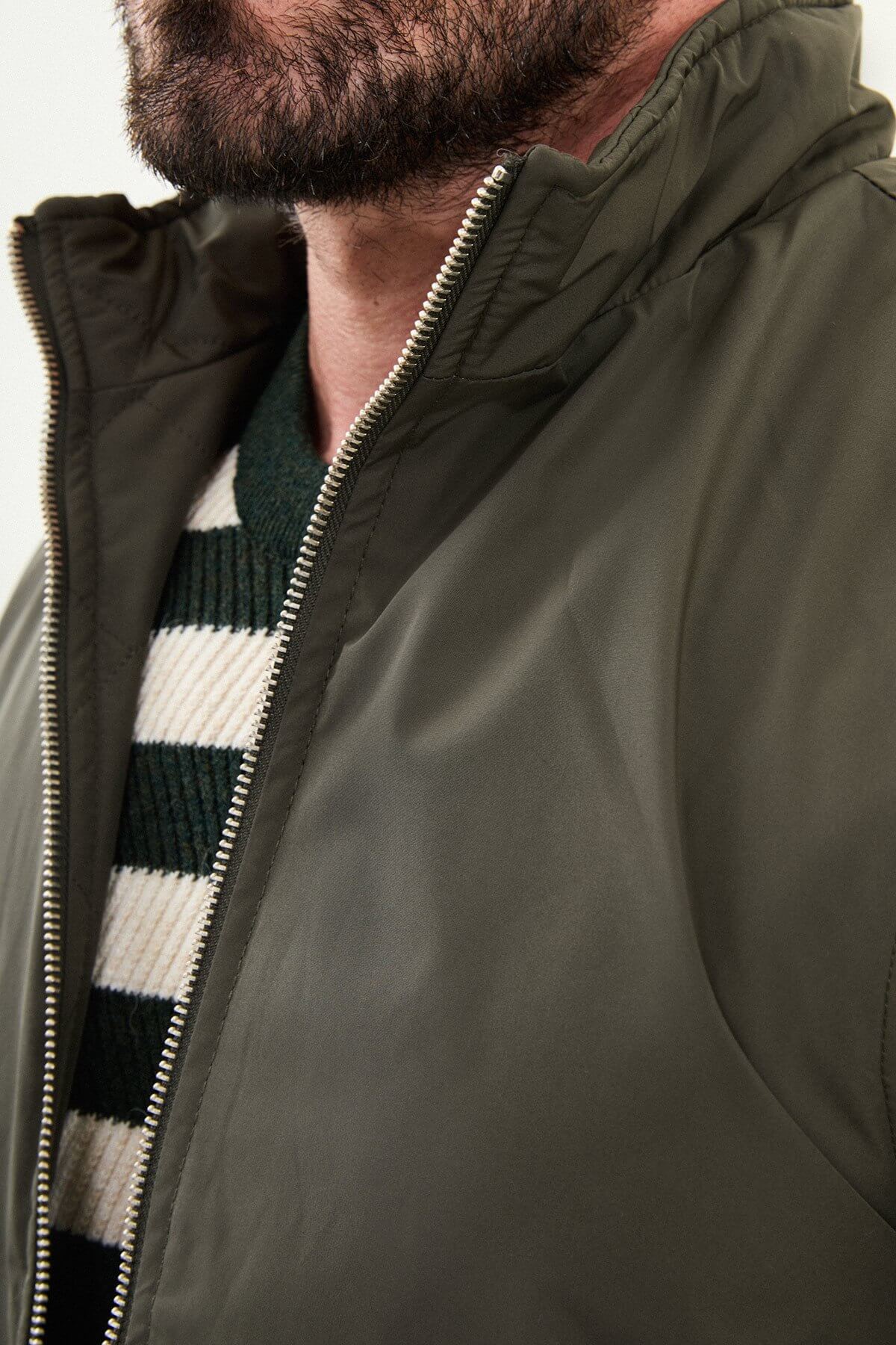 Men’s matte khaki/olive technical vest with a stand collar, zip front, and utility pockets, styled over a striped sweater for winter layering.