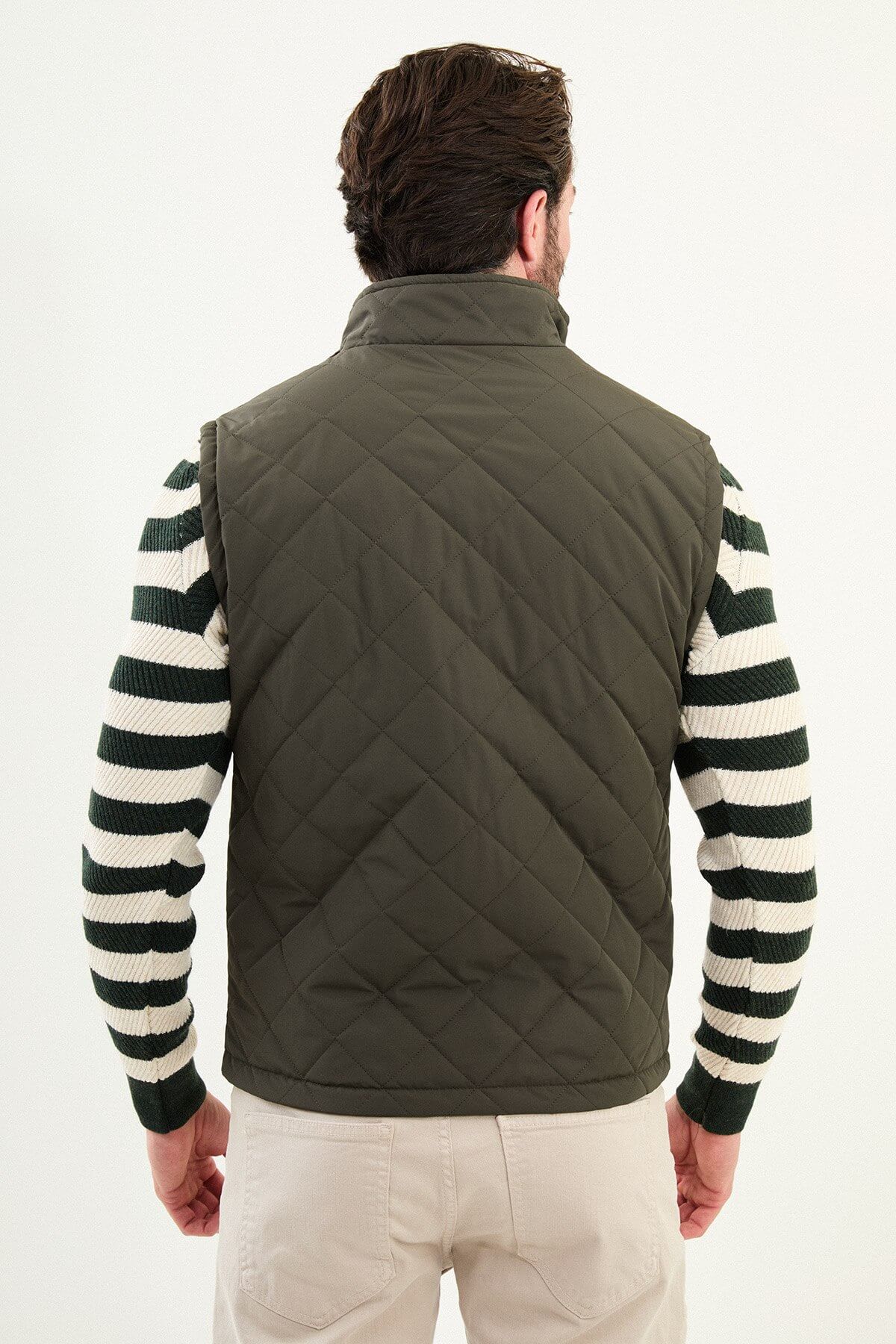 Men’s matte khaki/olive technical vest with a stand collar, zip front, and utility pockets, styled over a striped sweater for winter layering.